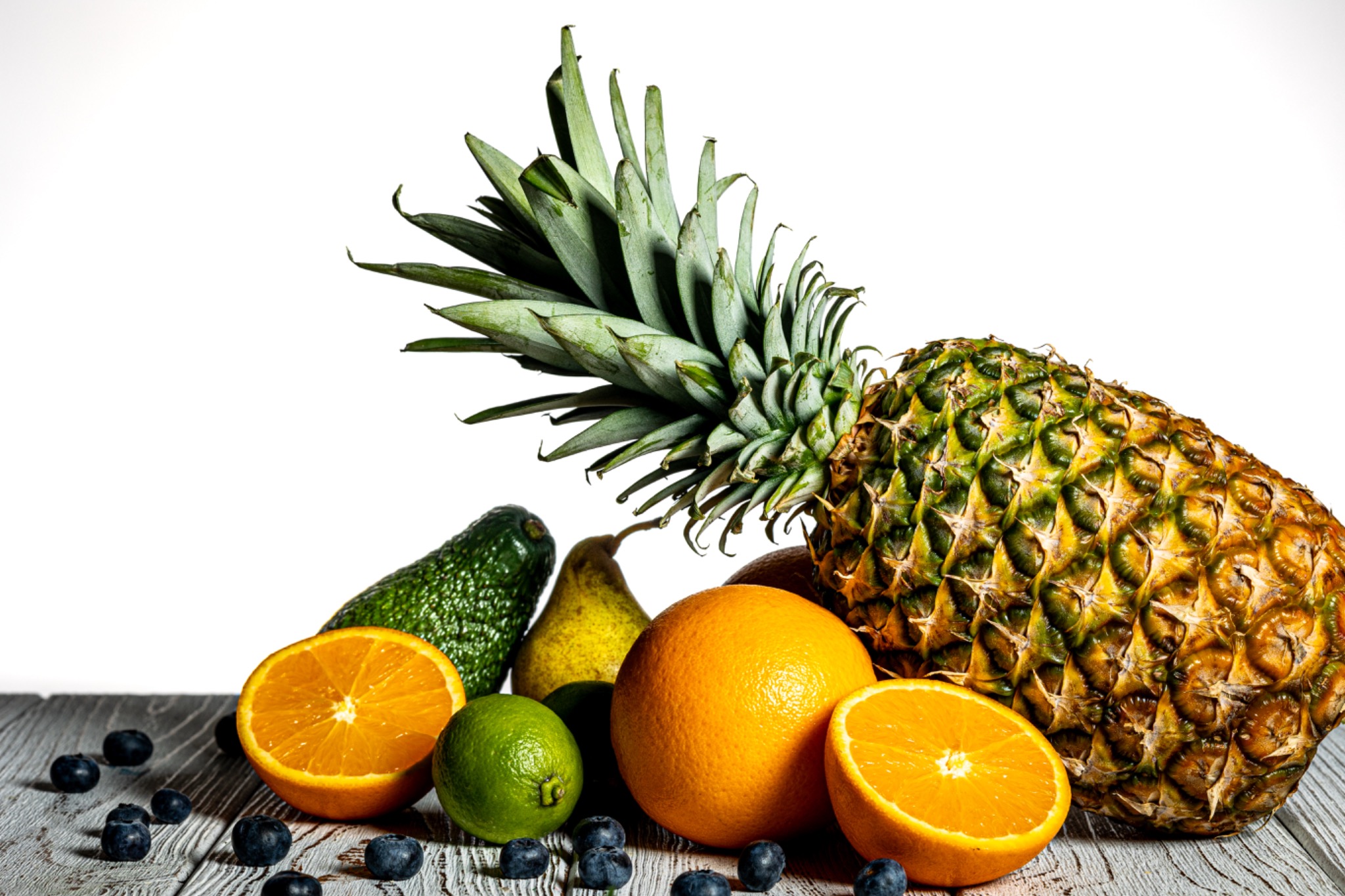 Martwa natura: ananas, pomarańcze (jedna przekrojona), limonka, awokado, gruszka i borówki na drewnianym blacie, białe tło. Owoce o intensywnych barwach.