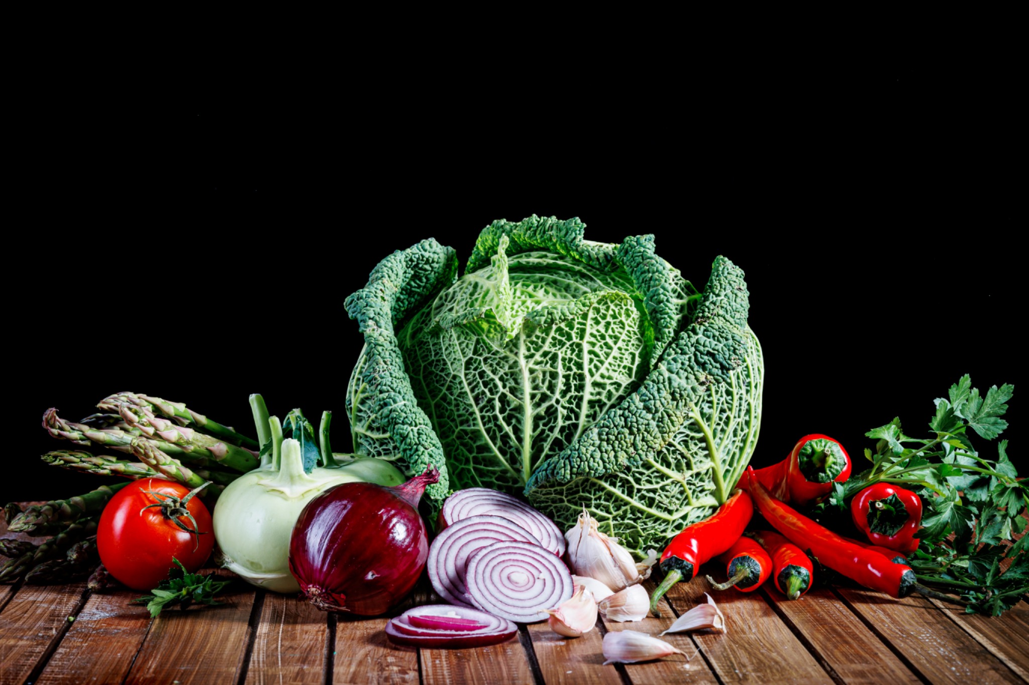 Martwa natura z warzyw na drewnianym stole: kapusta włoska, szparagi, pomidor, cebula czerwona, czosnek i papryka chili na czarnym tle. Zdjęcie w ciemnej tonacji.