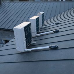 Dar-Max Artsem Churykau - Trzy białe jednostki klimatyzacji zamontowane na ciemnym, metalowym dachu domu. Widoczne przewody i elementy mocujące. W tle niebo o zmierzchu i zabudowa.