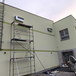 Dar-Max Artsem Churykau - Montaż klimatyzacji na elewacji budynku. Widoczne rusztowanie, drabina i dwa zamontowane urządzenia. Budynek w kolorze jasnożółtym z zielonym pasem. Częściowo widoczny samochód.