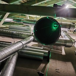 Dar-Max Artsem Churykau - Montaż instalacji wentylacyjnej w budynku. Widoczne rury wentylacyjne, poziomica laserowa i wkrętarka. Ujęcie z dołu, widoczna konstrukcja dachu.