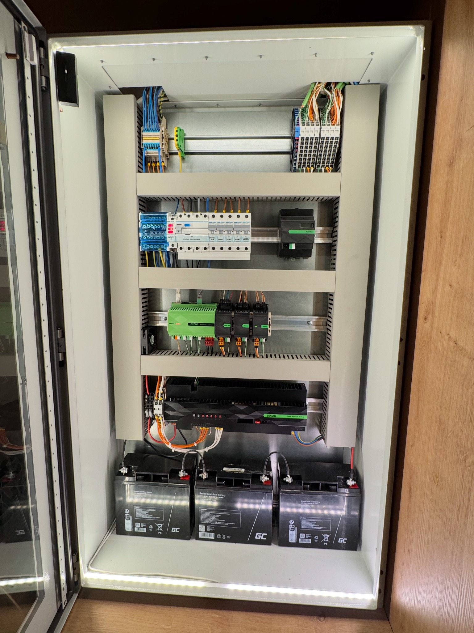 Rozdzielnica elektryczna z modułami Loxone i akumulatorami GC w obudowie z oświetleniem LED. Widoczne przewody i elementy sterujące. System inteligentnego domu.
