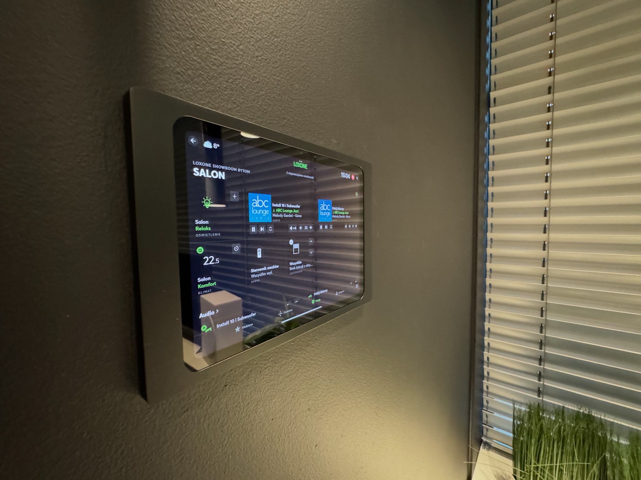 Panel sterowania inteligentnym domem Loxone w salonie, z widocznym interfejsem oświetlenia, klimatu i audio. Nowoczesny design, żaluzje w tle.