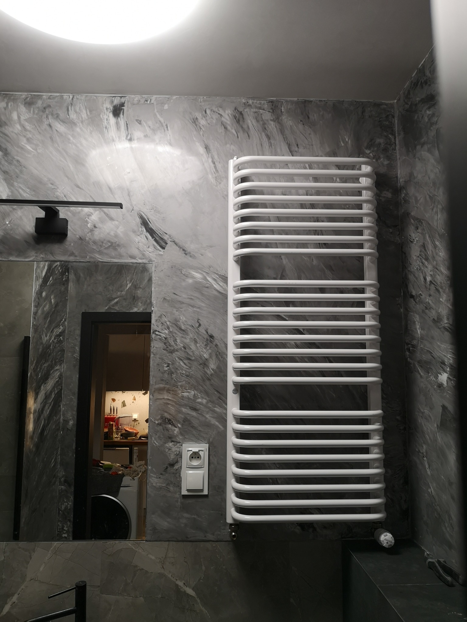 Biały grzejnik drabinkowy na tle ściany z efektem marmuru w szarościach. Widoczne lustro, lampa nad nim i fragment łazienki w odbiciu. Detal gniazdka elektrycznego.