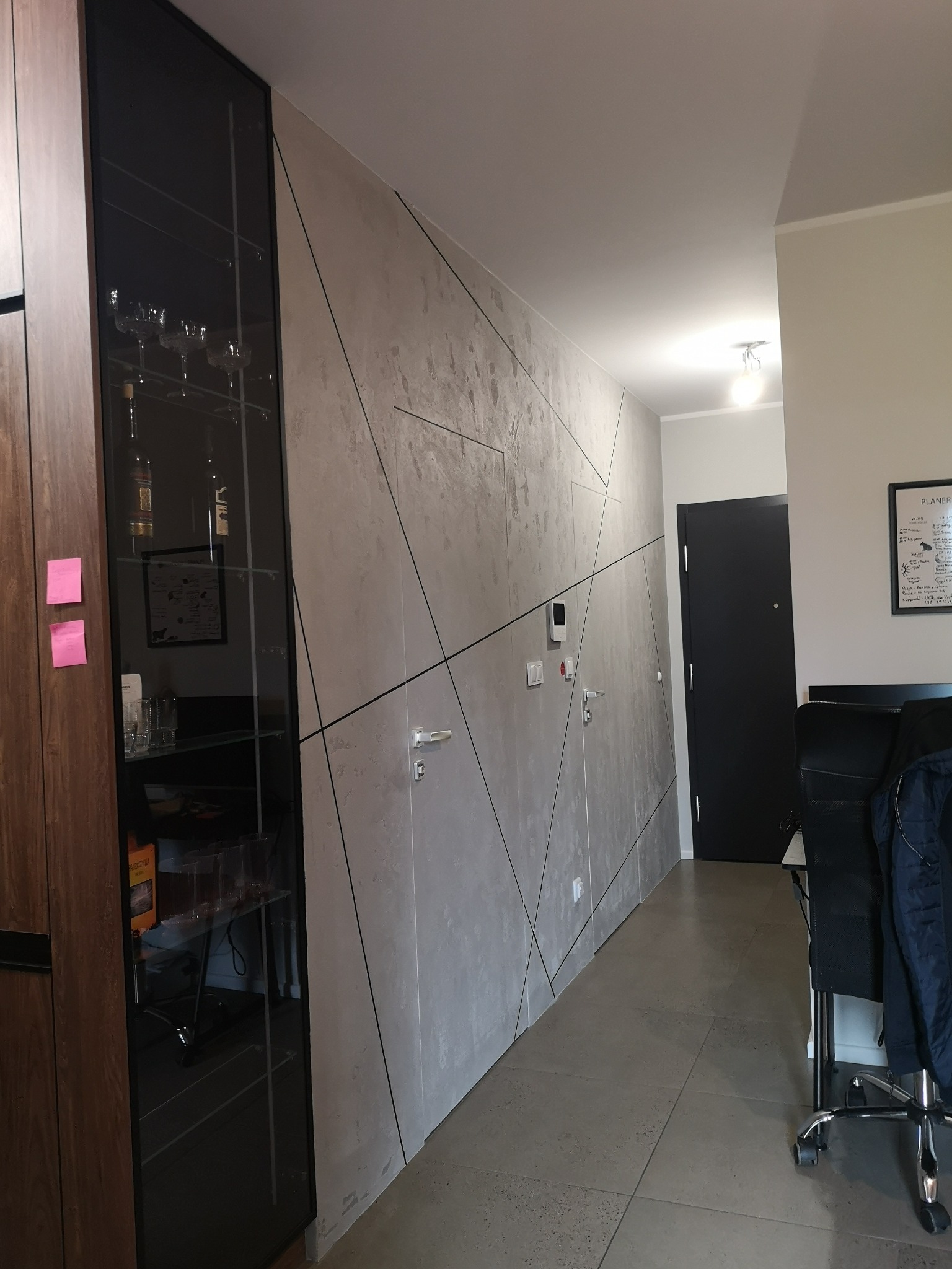 Nowoczesny korytarz z szarą ścianą o geometrycznym wzorze, ciemnymi drzwiami i częściowo widoczną przeszkloną szafką. Minimalistyczny design, gładka podłoga.