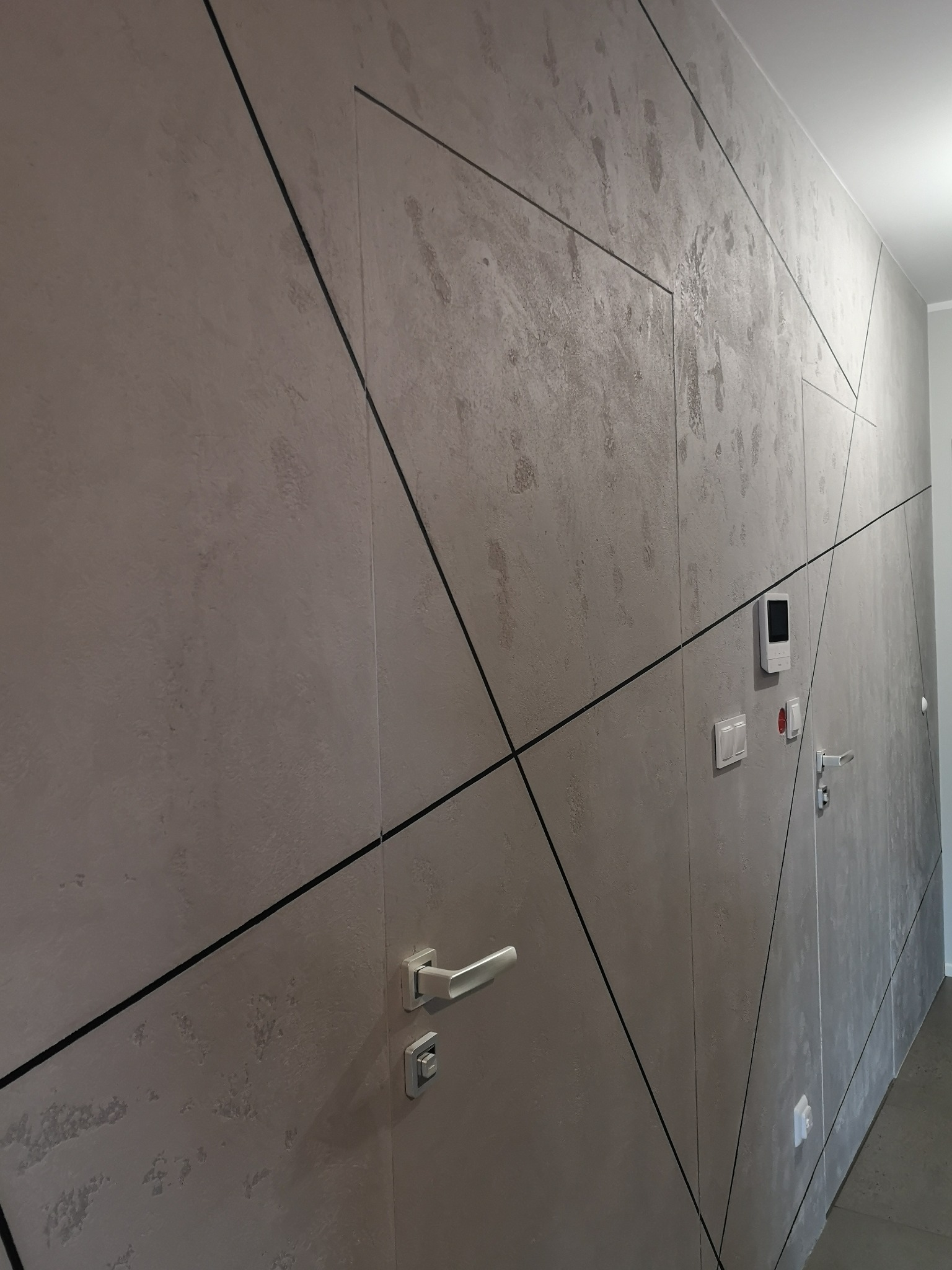 Ściana z ukrytymi drzwiami, wykończona szarą, strukturalną farbą z czarnymi, geometrycznymi liniami. Widoczne białe klamki, włączniki i panel sterowania.