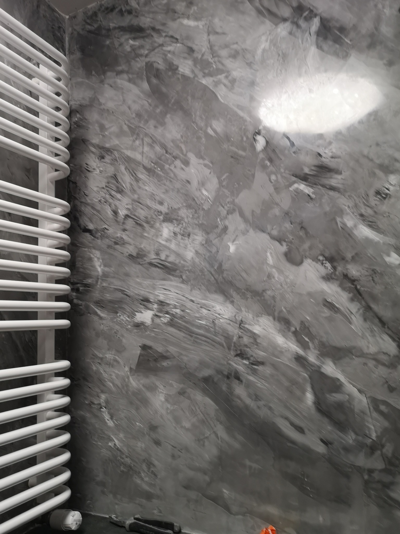 Dekoracyjna ściana w odcieniach szarości z efektem betonu w łazience, obok biały grzejnik drabinkowy. Widoczne narzędzia malarskie na podłodze.