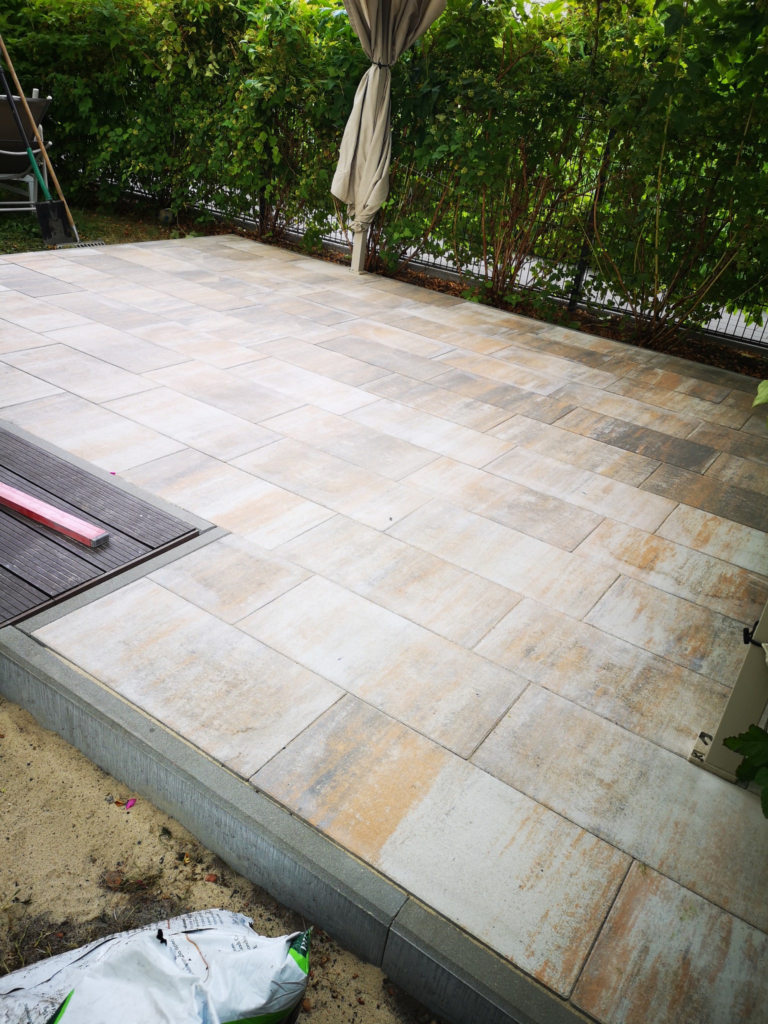 Wyłożony kostką taras z elementami drewnianymi i obrzeżem z kostki betonowej. W tle zielony żywopłot i częściowo złożony parasol.