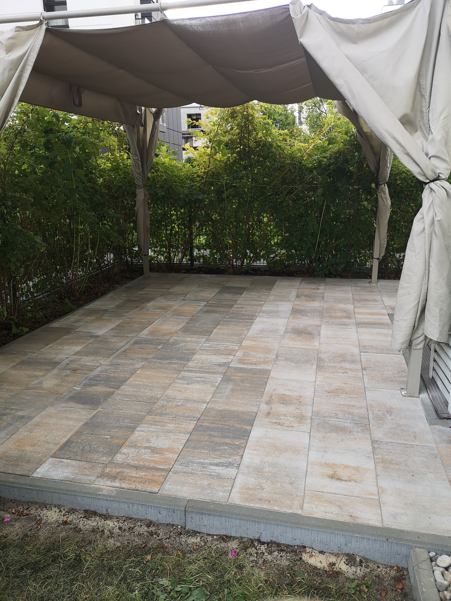 Nowoczesny, wybrukowany taras z zadaszeniem, otoczony zielenią. Widoczne detale kostki brukowej i starannie wykonane obrzeże.