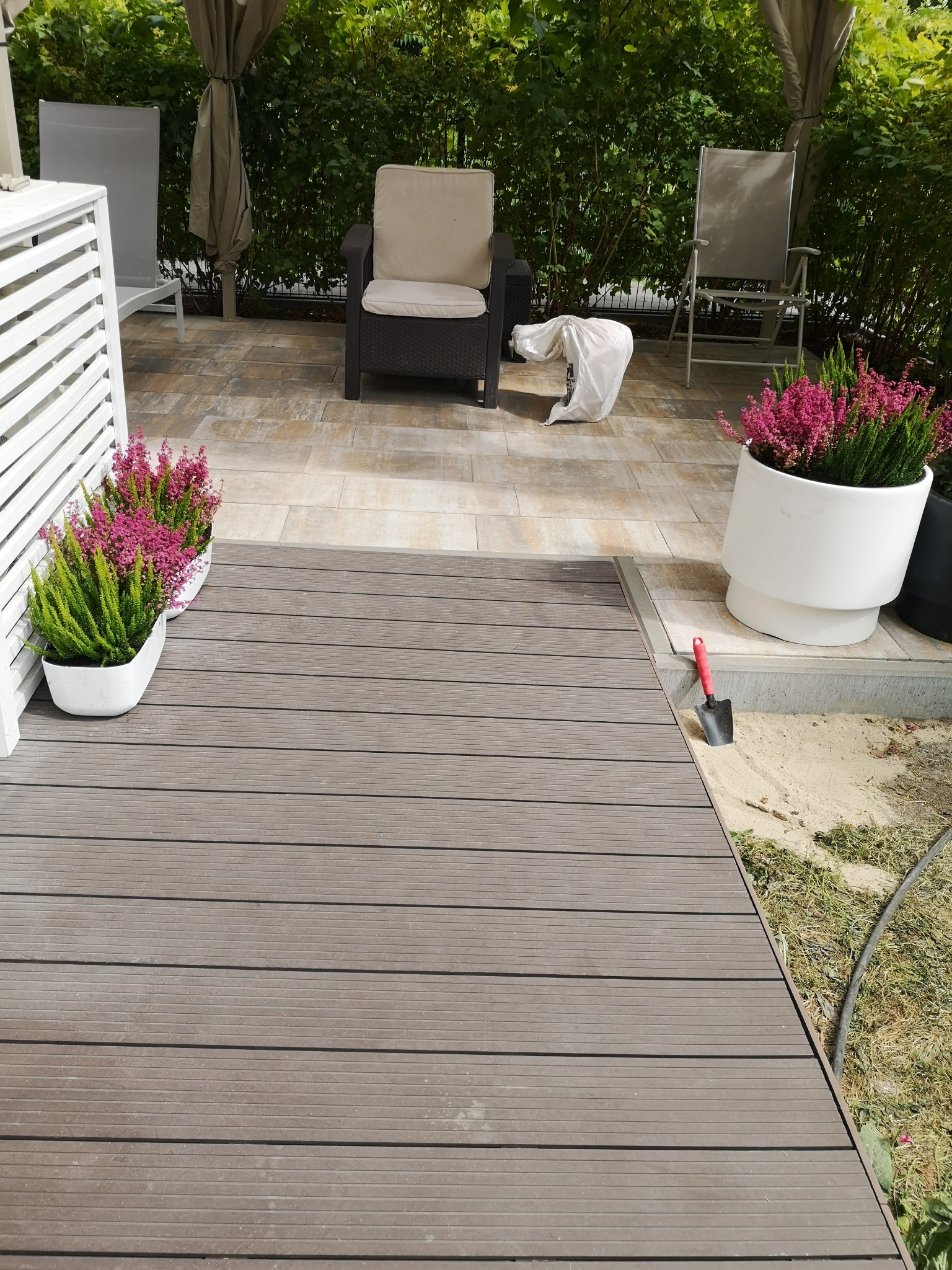 Nowy taras z desek kompozytowych w kolorze brązu, z donicami z wrzosami, meblami ogrodowymi i płytkami. Widoczna mała łopatka ogrodowa.