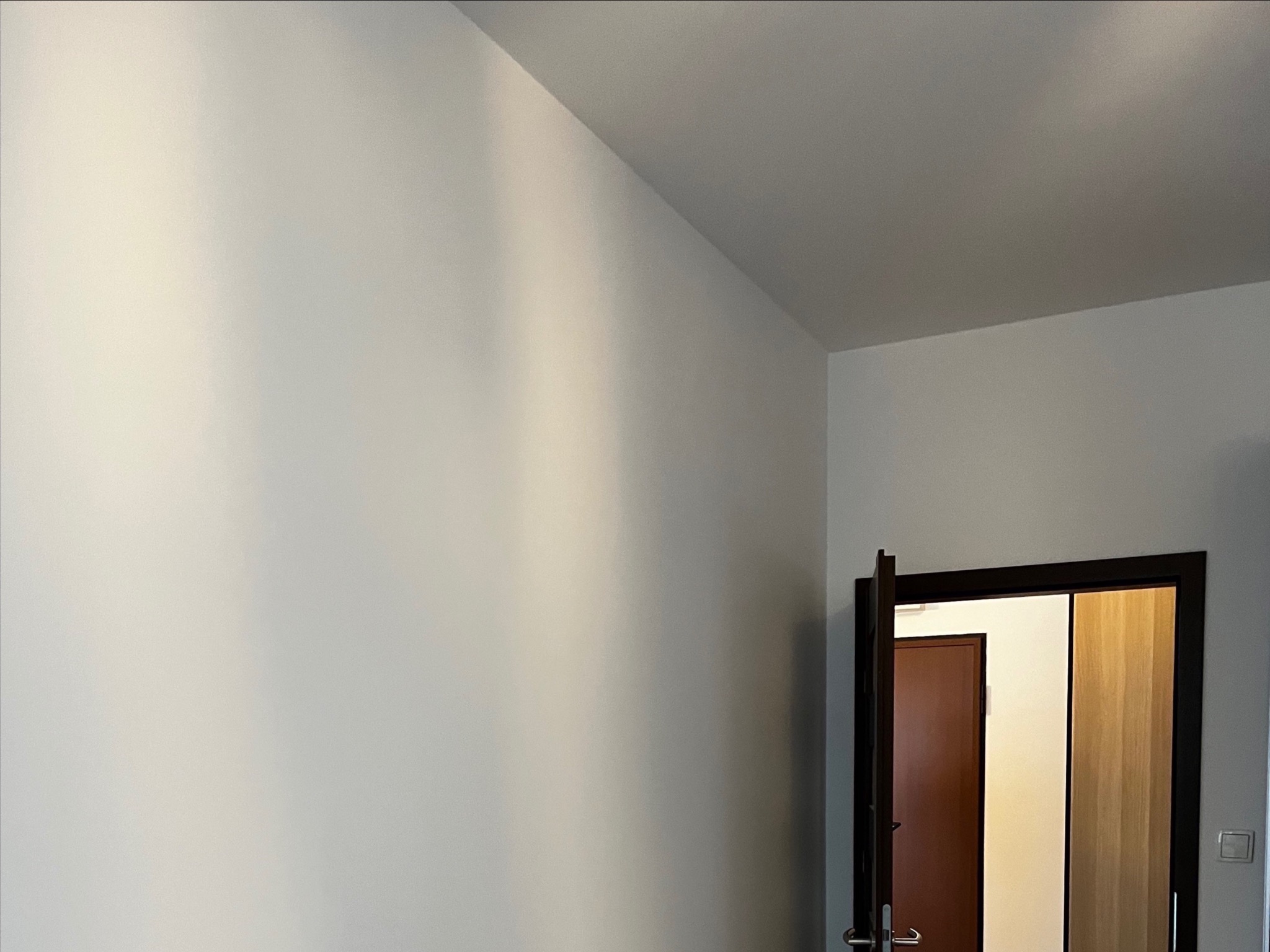 Jasna ściana po szpachlowaniu i malowaniu, widoczny fragment otwartych drzwi z ciemną ościeżnicą. Minimalistyczne wnętrze po remoncie.