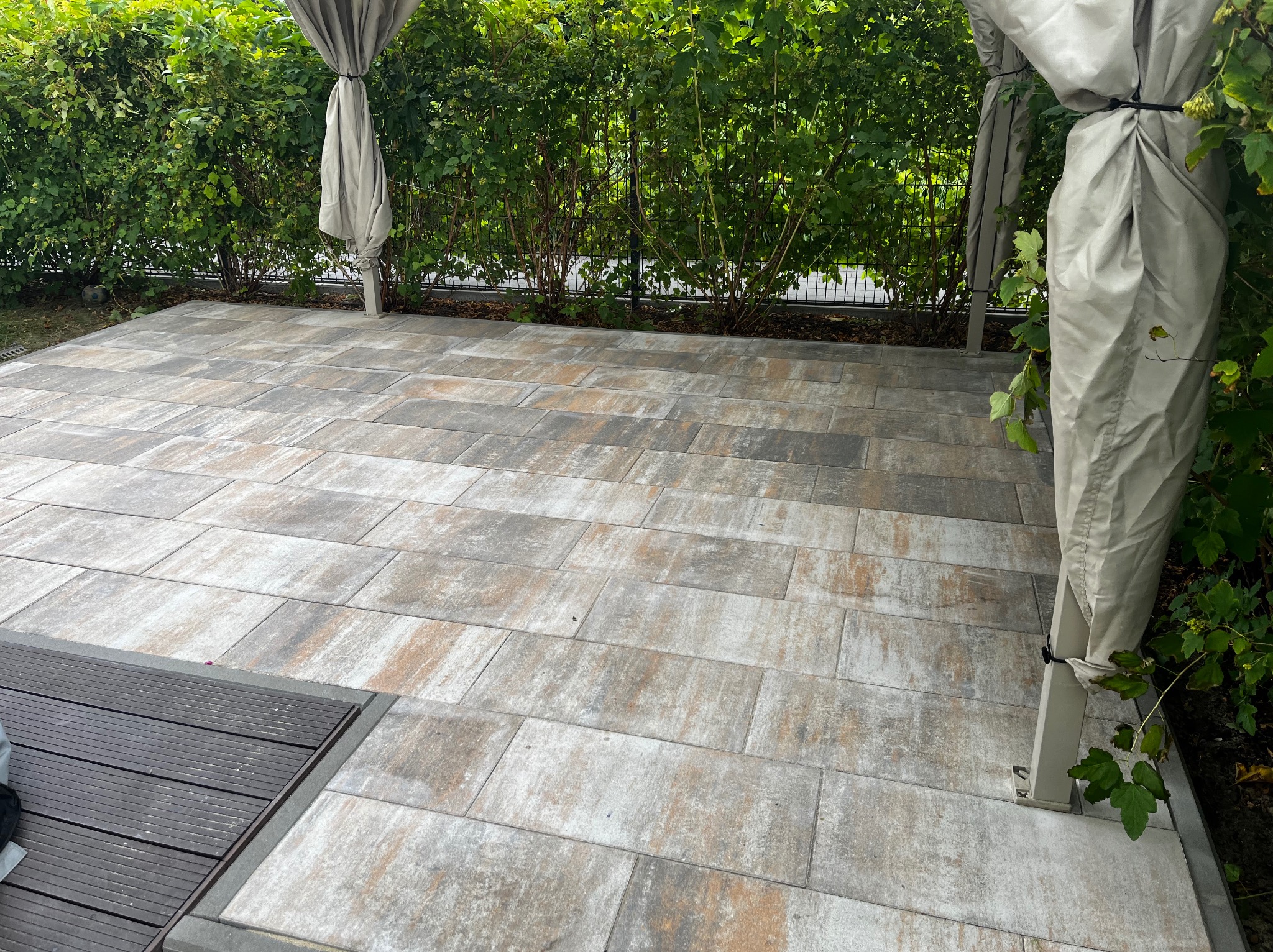 Ułożona, wielobarwna kostka brukowa na tarasie z drewnianym podestem, otoczona zielenią i elementami konstrukcji z zasłonami. Widok na zewnątrz.