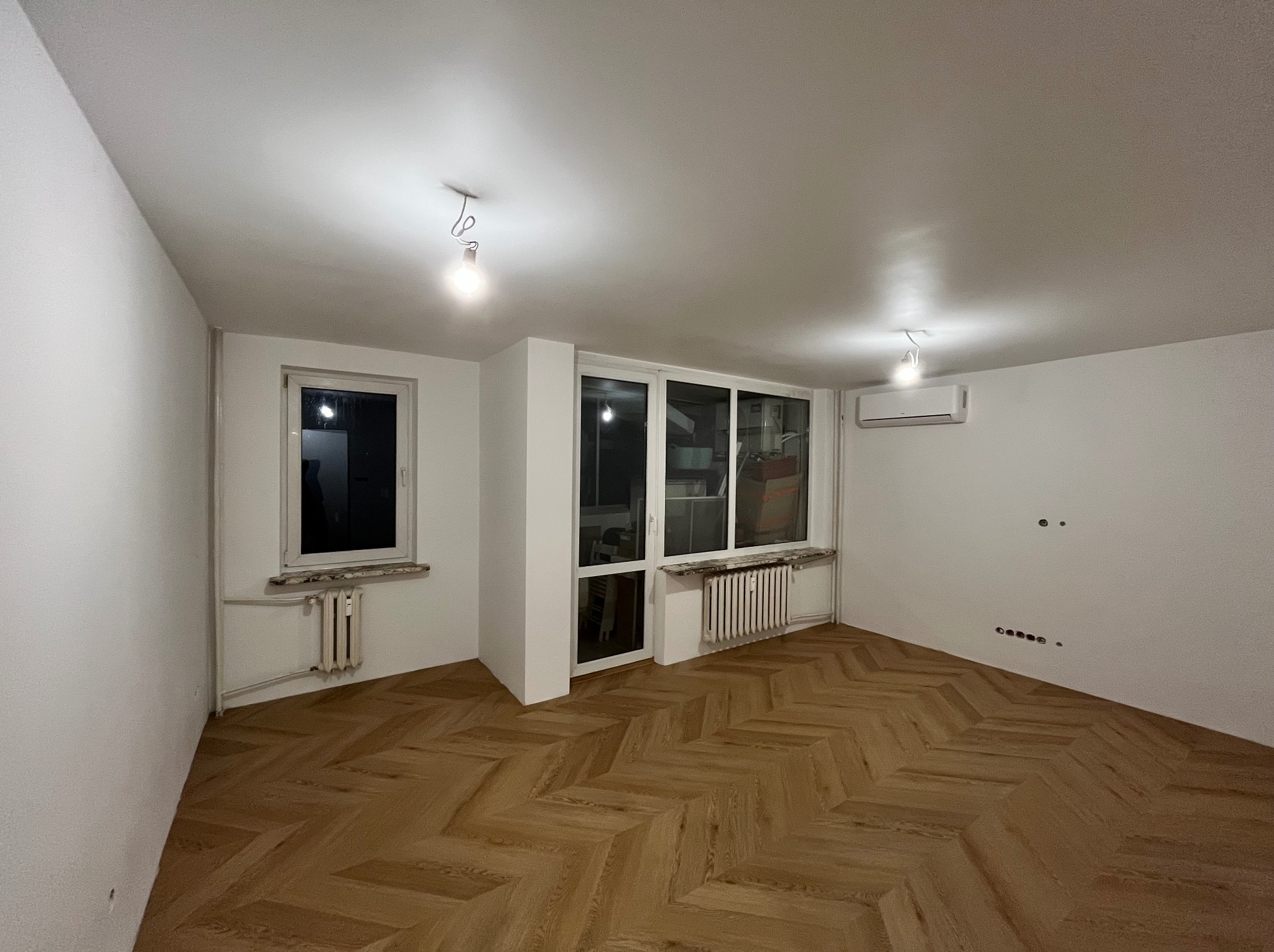 Jasne, puste wnętrze po remoncie: parkiet w jodełkę, białe ściany, okno i balkon. Widoczne grzejniki, klimatyzacja i punkty elektryczne. Minimalistyczny design.