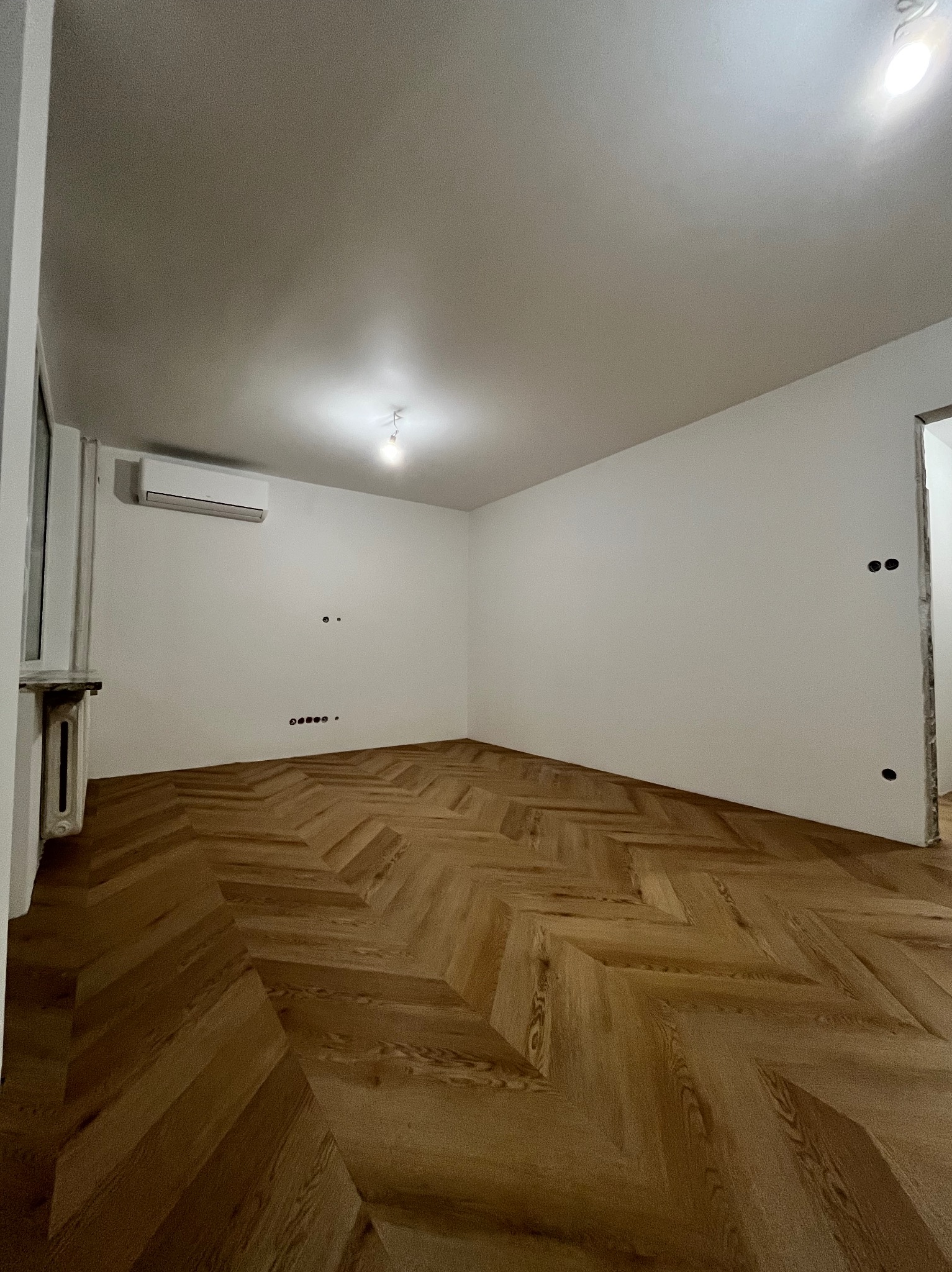 Jasne, puste pomieszczenie po remoncie z parkietem ułożonym w jodełkę. Widoczne gładkie, białe ściany, klimatyzacja i otwór drzwiowy w stanie surowym.