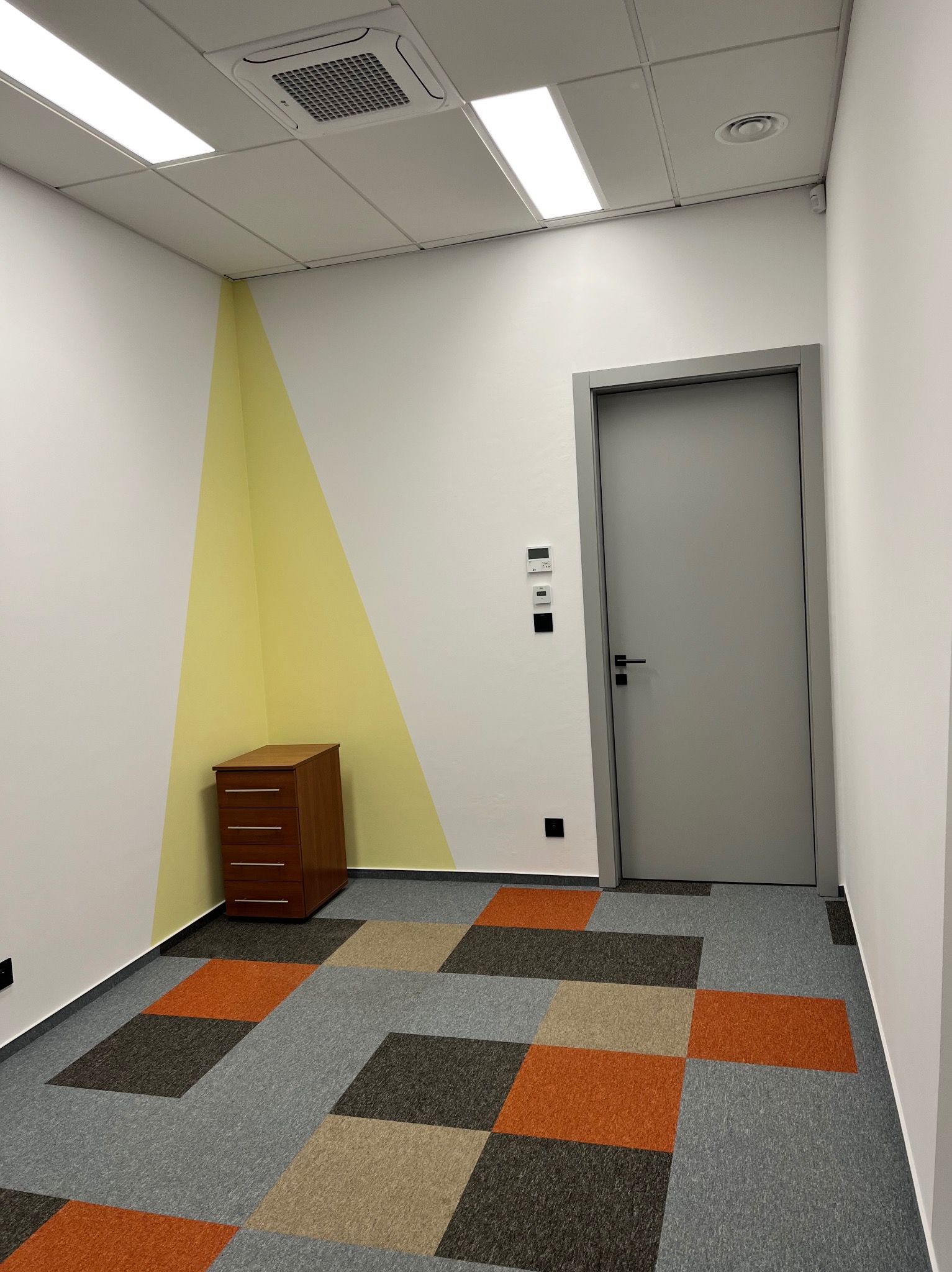 Nowoczesne wykończenie biura w Warszawie: szara stolarka drzwiowa, geometryczne malowanie ścian w odcieniach żółci i brązu, oraz wzorzysta wykładzina podłogowa.