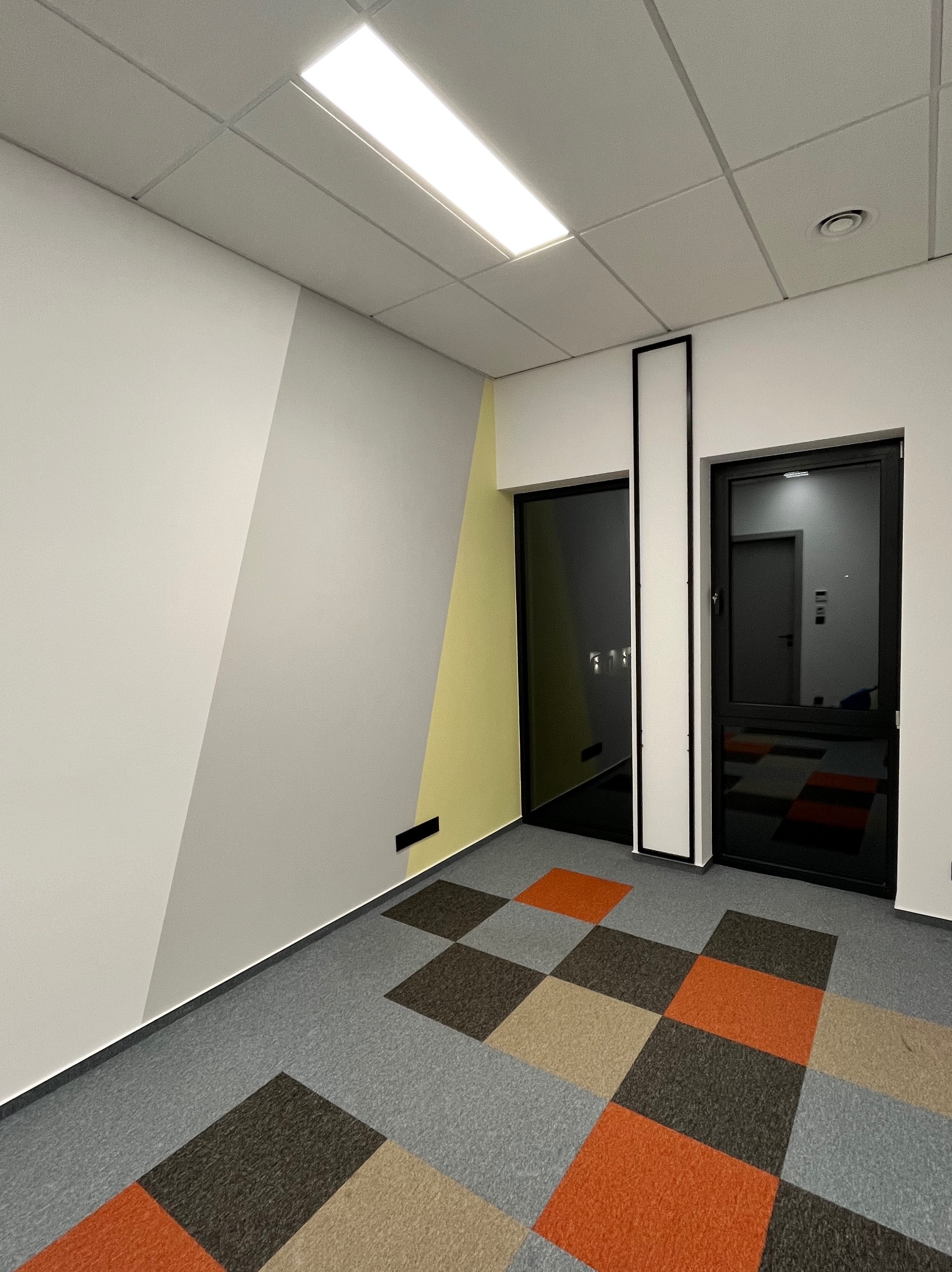 Nowoczesne wnętrze biurowe po remoncie: geometryczne malowanie ścian w odcieniach szarości i żółci, wzorzysta wykładzina podłogowa, czarne ramy drzwi.