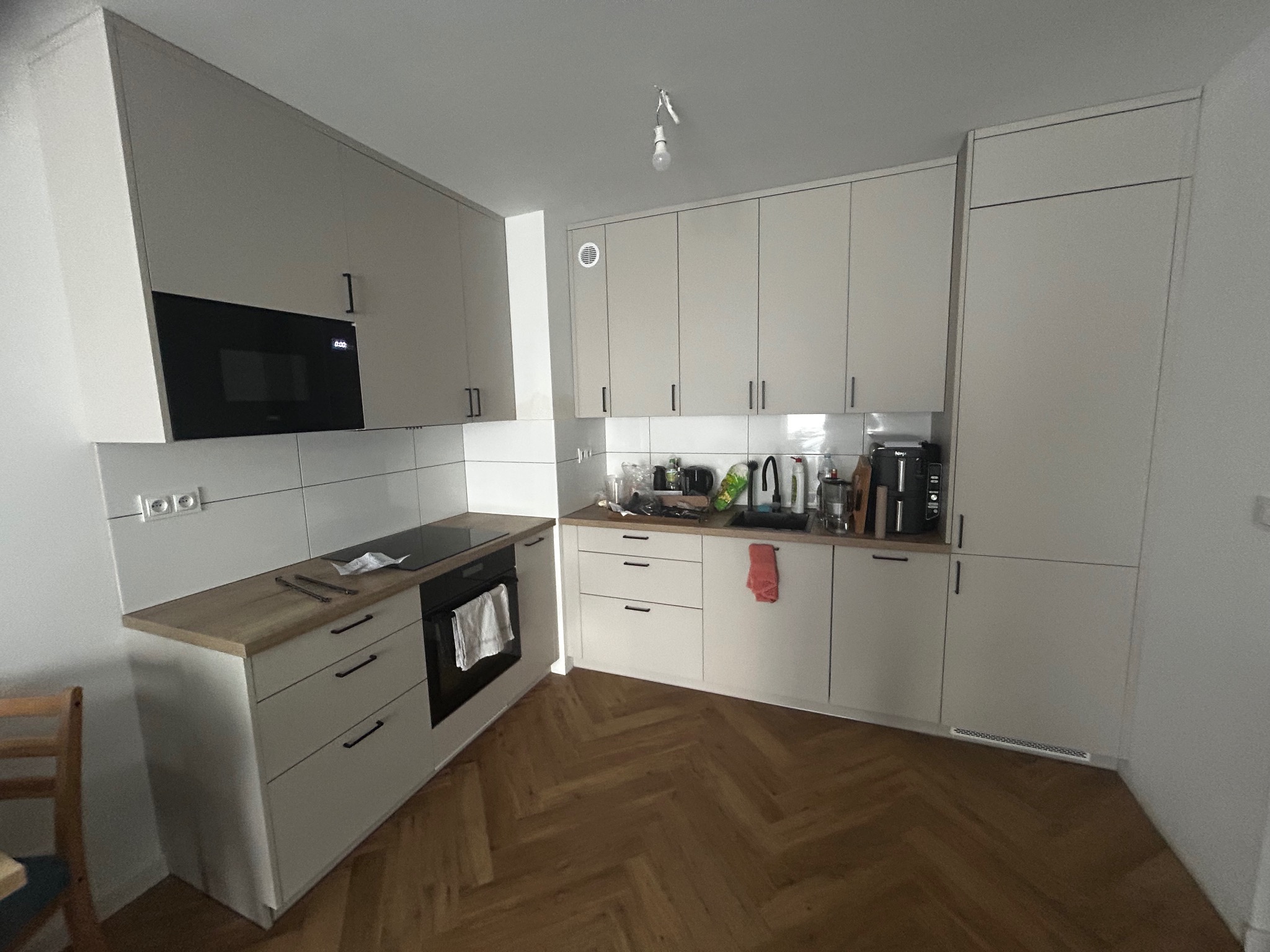 Jasna kuchnia z drewnianym blatem i parkietem w jodełkę. Zabudowa kuchenna z czarnymi uchwytami, mikrofalówka w zabudowie. Widoczny fragment stołu i krzesła.