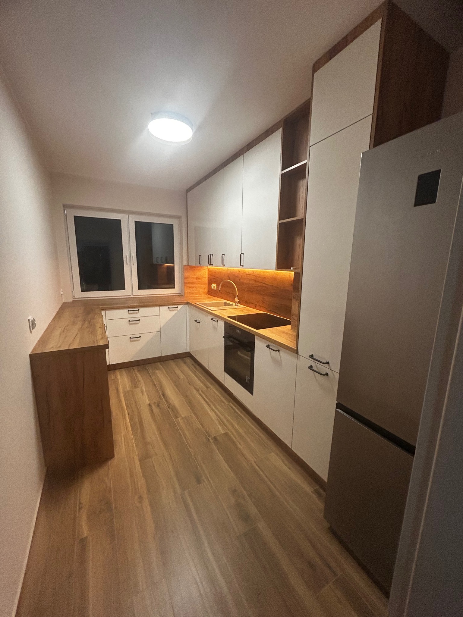 Nowoczesna, jasna kuchnia z drewnianymi akcentami. Białe fronty szafek, drewniany blat i panele podłogowe. Oświetlenie LED pod szafkami. Widoczna lodówka.