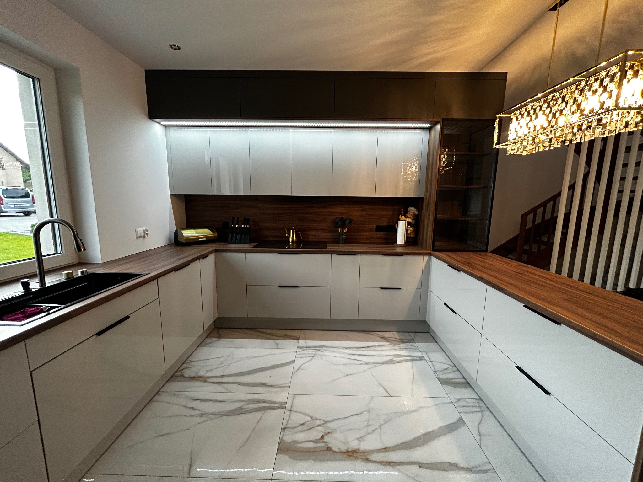 Nowoczesna kuchnia w kształcie litery U z białymi, lakierowanymi frontami, drewnianym blatem i marmurową podłogą. Nad blatem rząd szafek z oświetleniem LED.