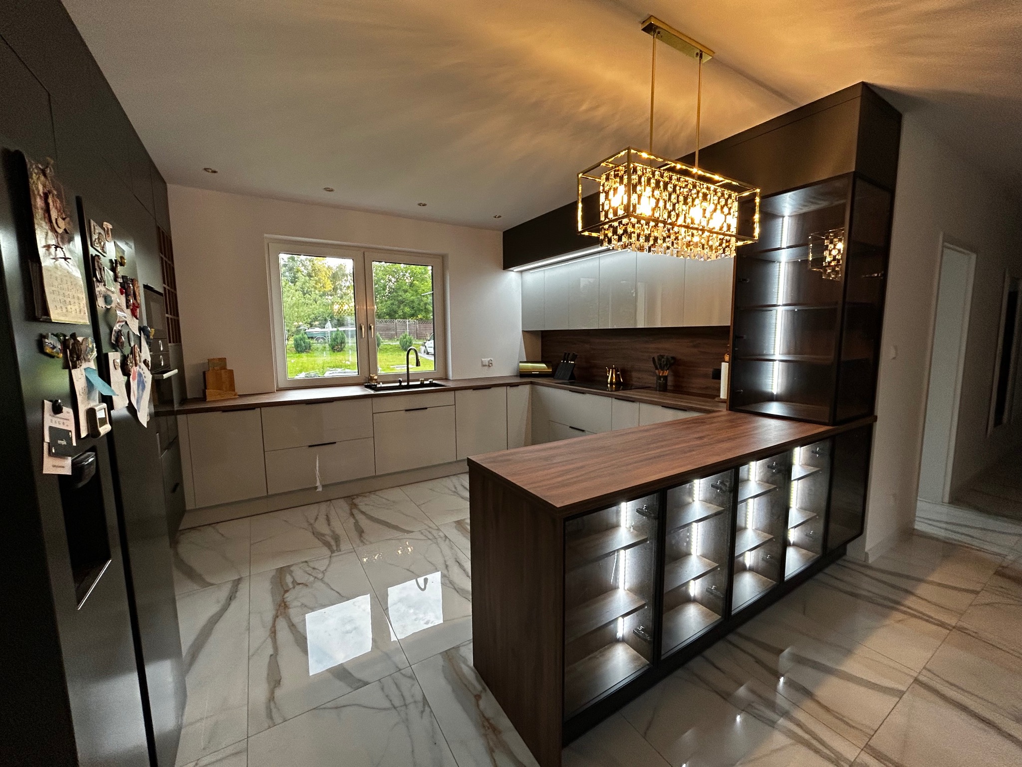 Nowoczesna kuchnia z wyspą i oświetleniem LED. Białe fronty szafek, drewniany blat. Kryształowy żyrandol. Marmurowe płytki podłogowe. Widok na ogród przez okno.