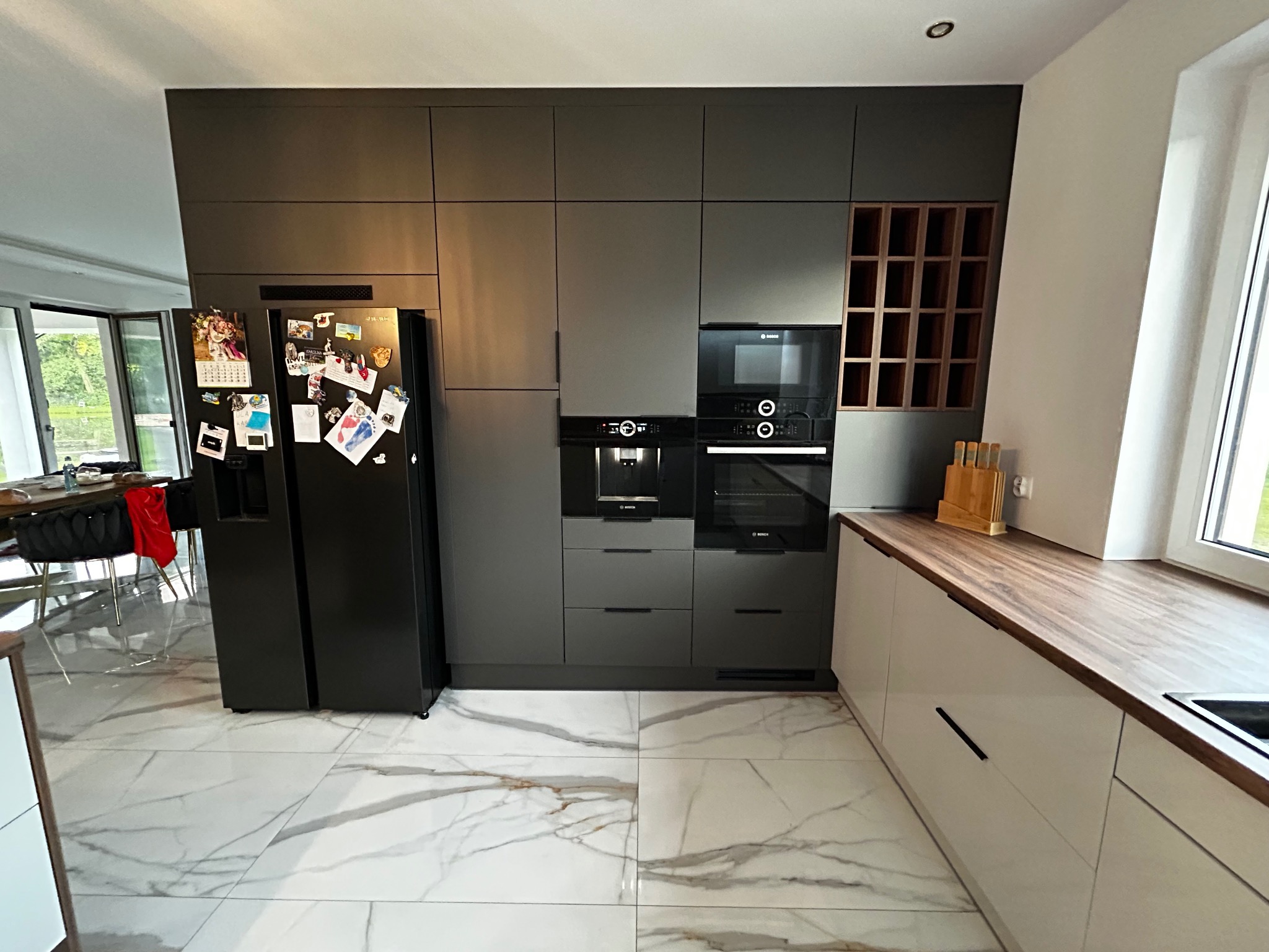 Nowoczesna kuchnia z szarymi frontami i drewnianym blatem. Na lodówce magnesy i zdjęcia. Marmurowe płytki podłogowe. Widoczny ekspres do kawy i piekarnik marki Bosch.