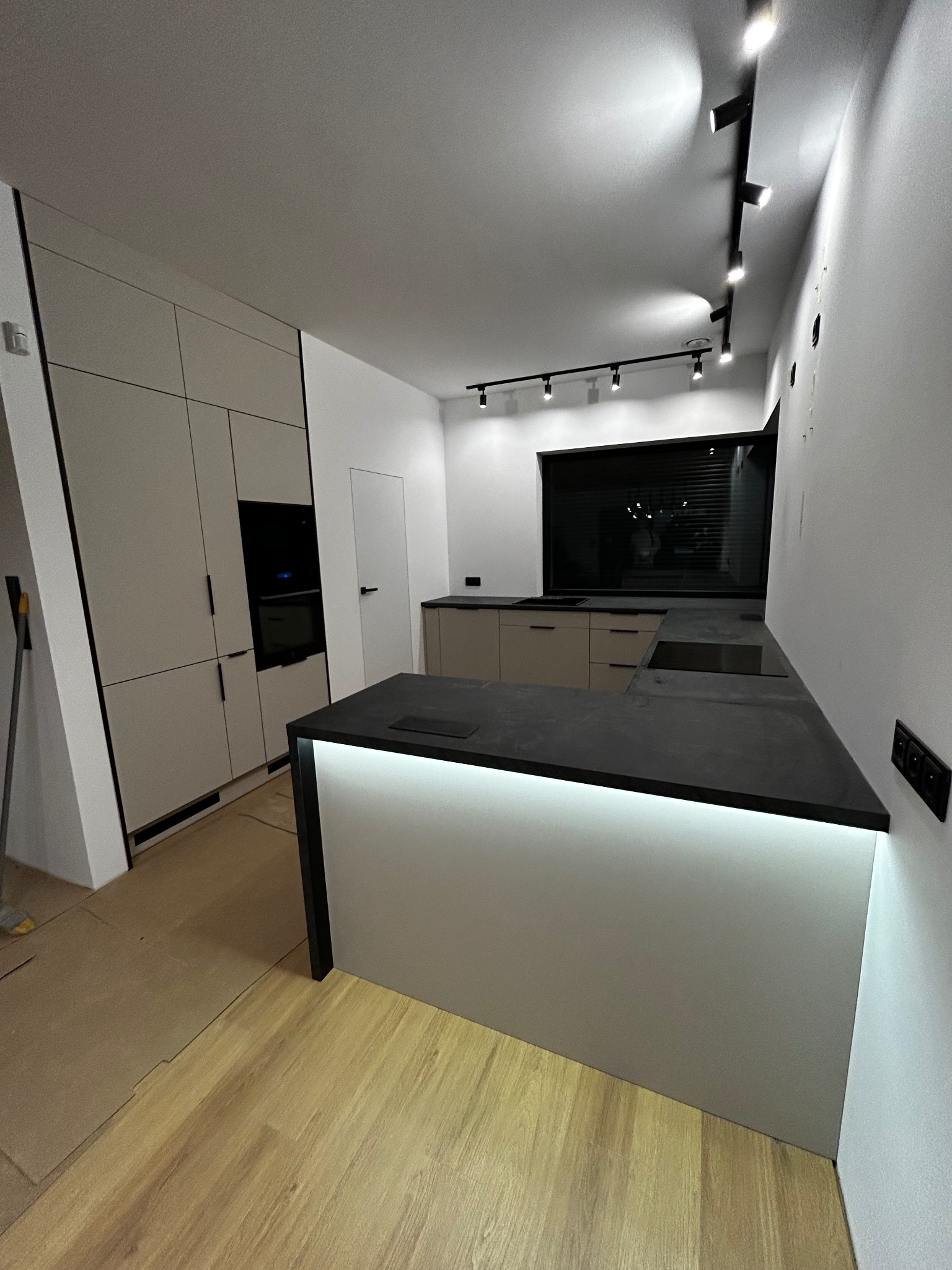 Nowoczesna kuchnia z wyspą i oświetleniem LED pod blatem. Minimalistyczny design z jasnymi frontami i czarnymi akcentami. Widoczne panele podłogowe i oświetlenie sufitowe.