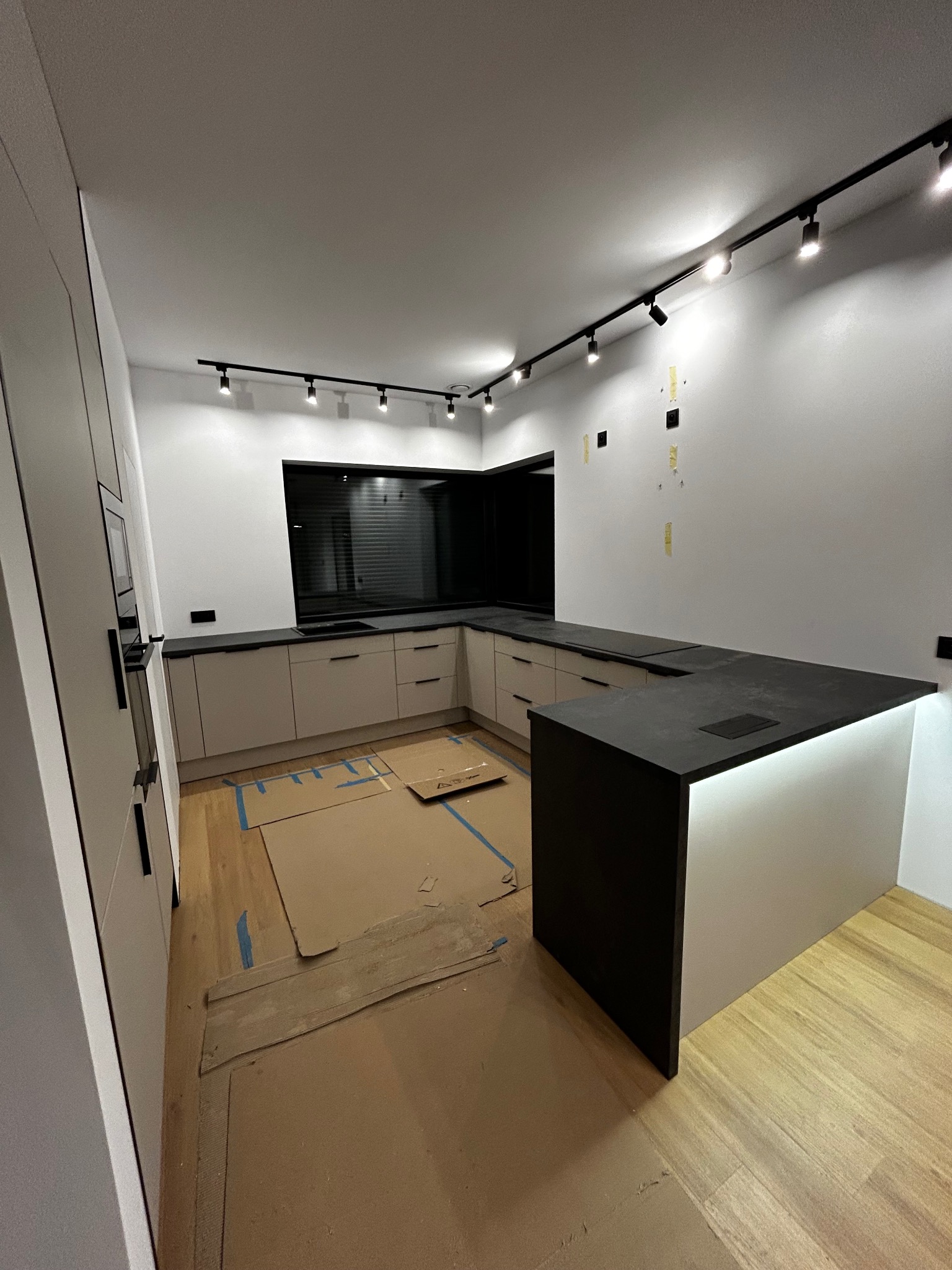 Nowoczesna, beżowa kuchnia z czarnym blatem i oświetleniem punktowym. Na podłodze kartony zabezpieczające. Widoczne gniazdka na ścianie.
