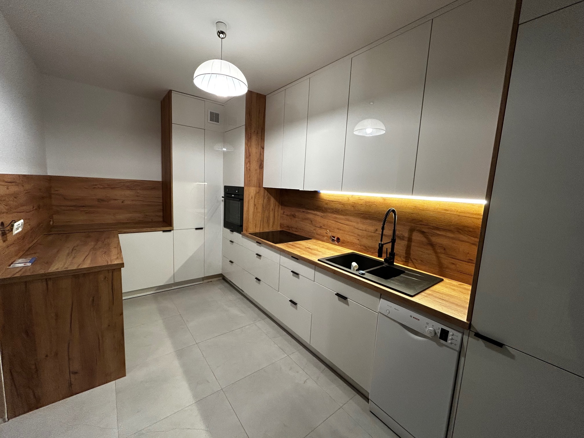 Nowoczesna kuchnia z białymi szafkami i drewnianym blatem. Oświetlenie LED podkreśla ciepło drewna. Czarny zlew i bateria dodają elegancji.