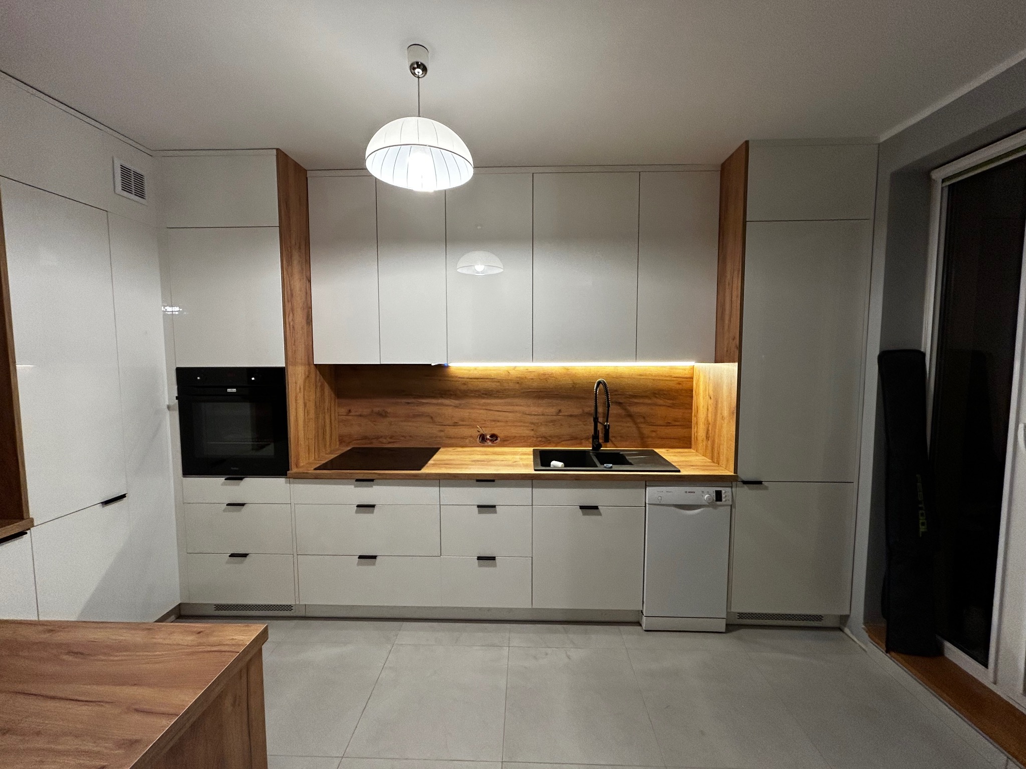 Nowoczesna kuchnia w bieli i drewnie w Rokietnicy. Szafki z minimalistycznymi uchwytami, drewniany blat i oświetlenie LED tworzą przytulną atmosferę.