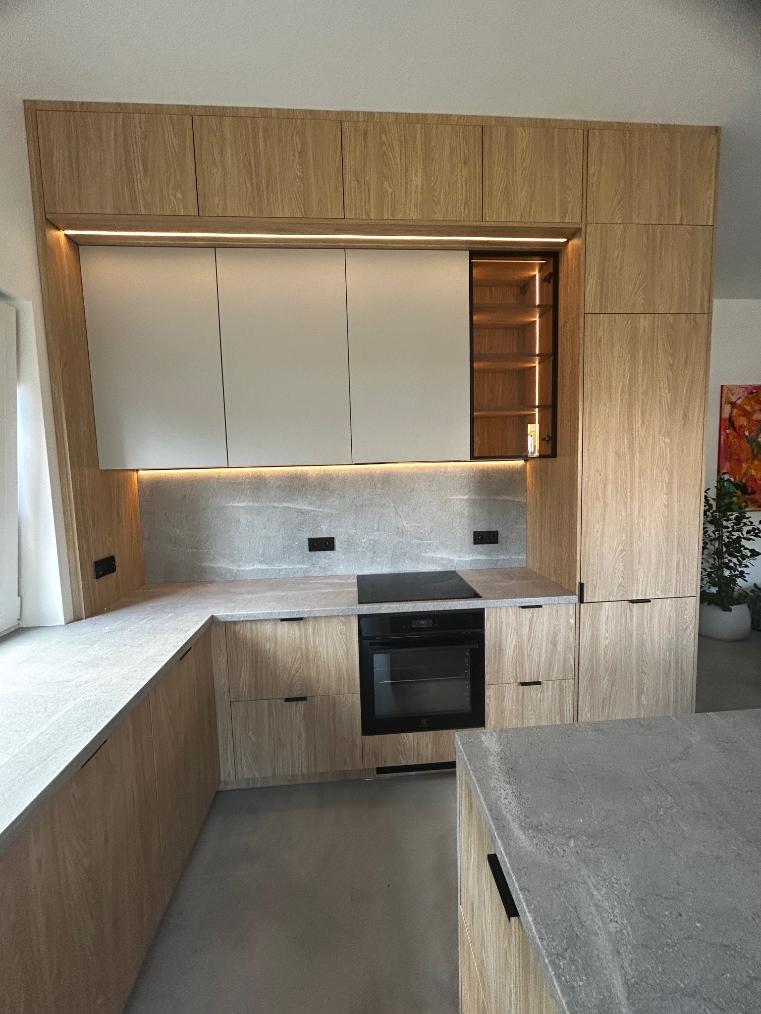 Nowoczesna kuchnia na wymiar z drewnianymi frontami, szarym blatem i oświetleniem LED. Widoczna zabudowa piekarnika i szafka z półkami podświetlonymi od wewnątrz.