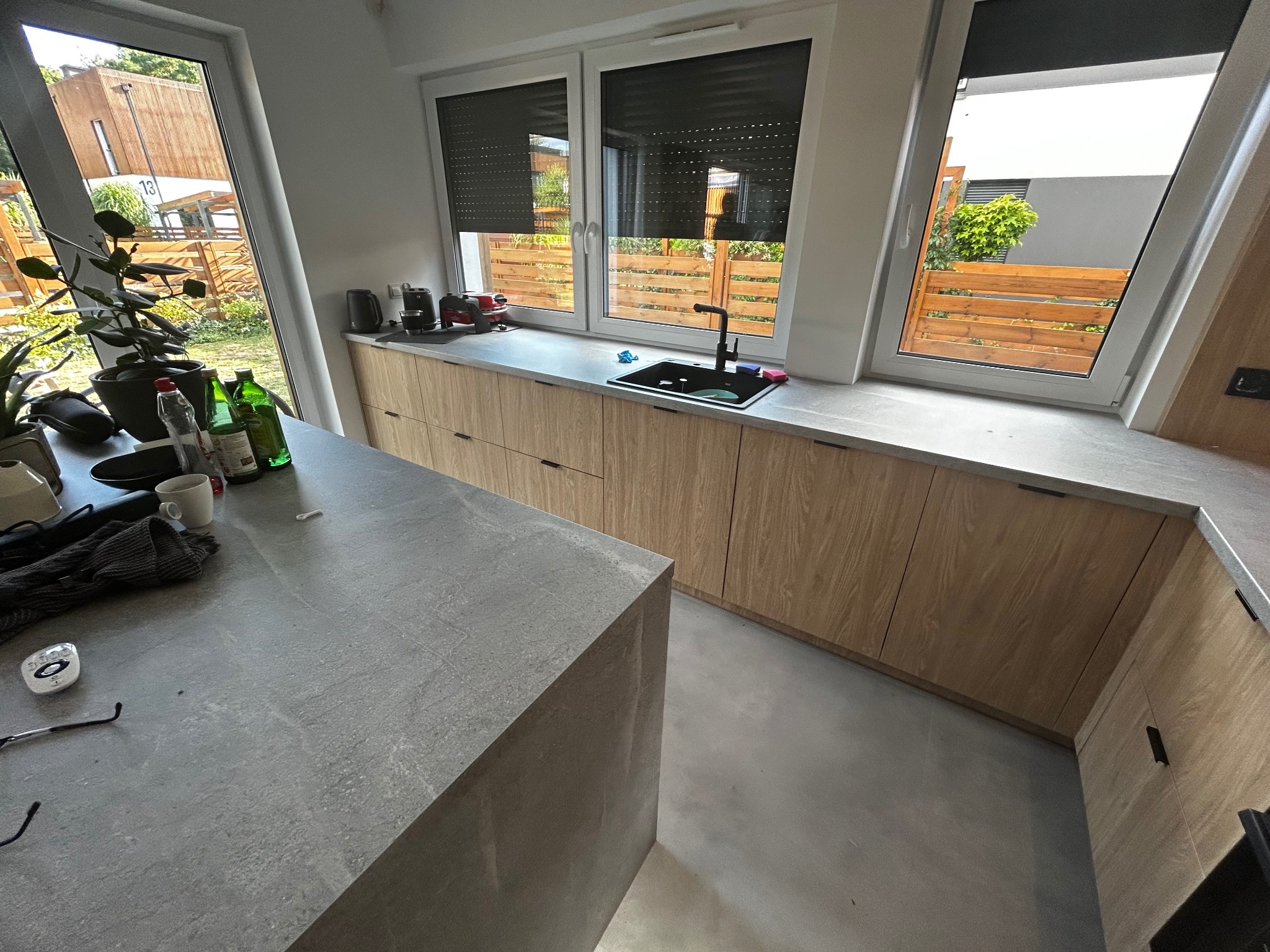 Jasna kuchnia z drewnianymi frontami i szarym blatem, widok na zewnątrz przez okno z roletami. Minimalistyczny design z wyspą kuchenną i elementami wyposażenia.