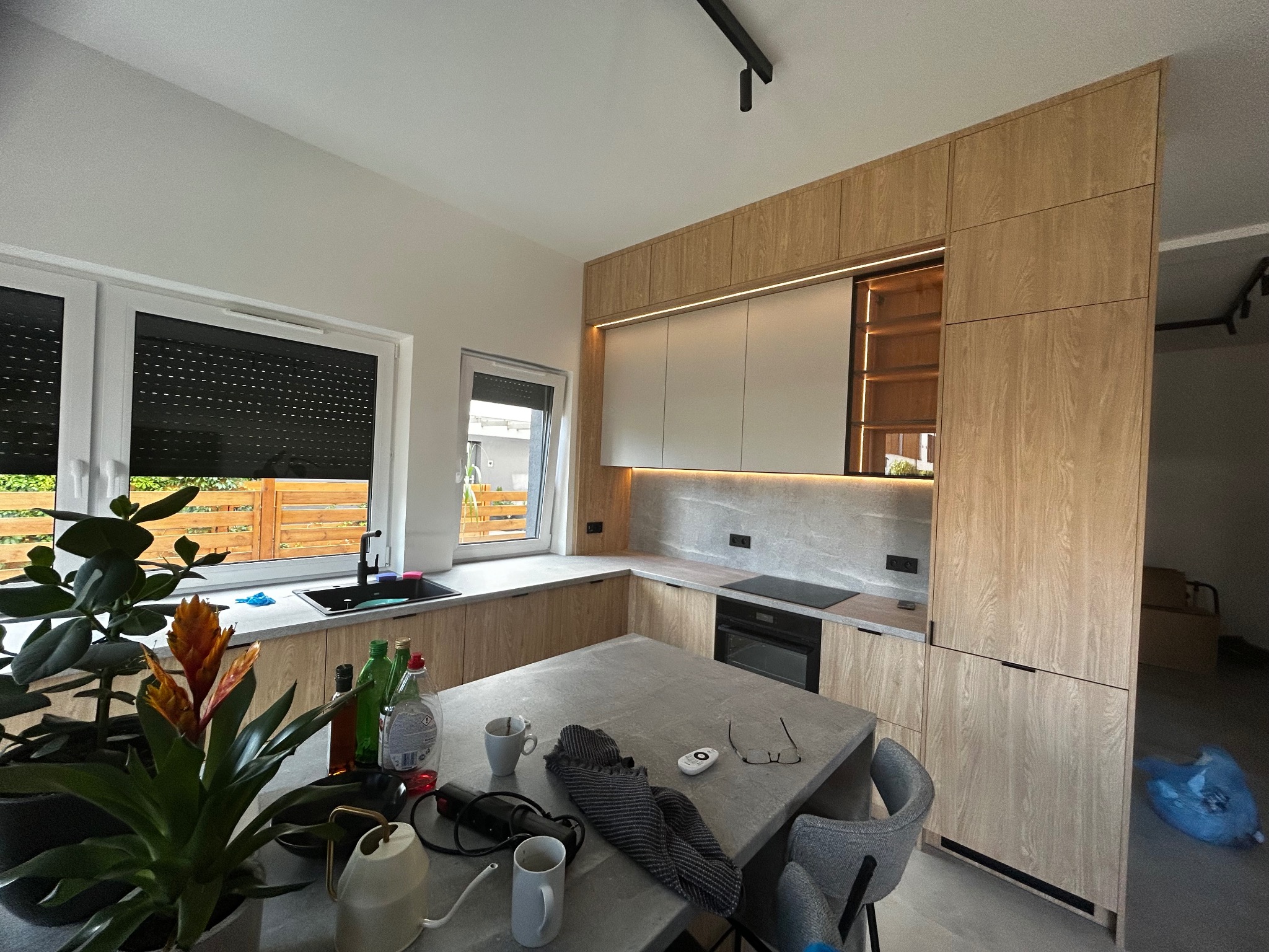 Nowoczesna kuchnia z drewnianymi frontami i szarym blatem. Na pierwszym planie roślina doniczkowa i akcesoria kuchenne. Ujęcie z perspektywy użytkownika.