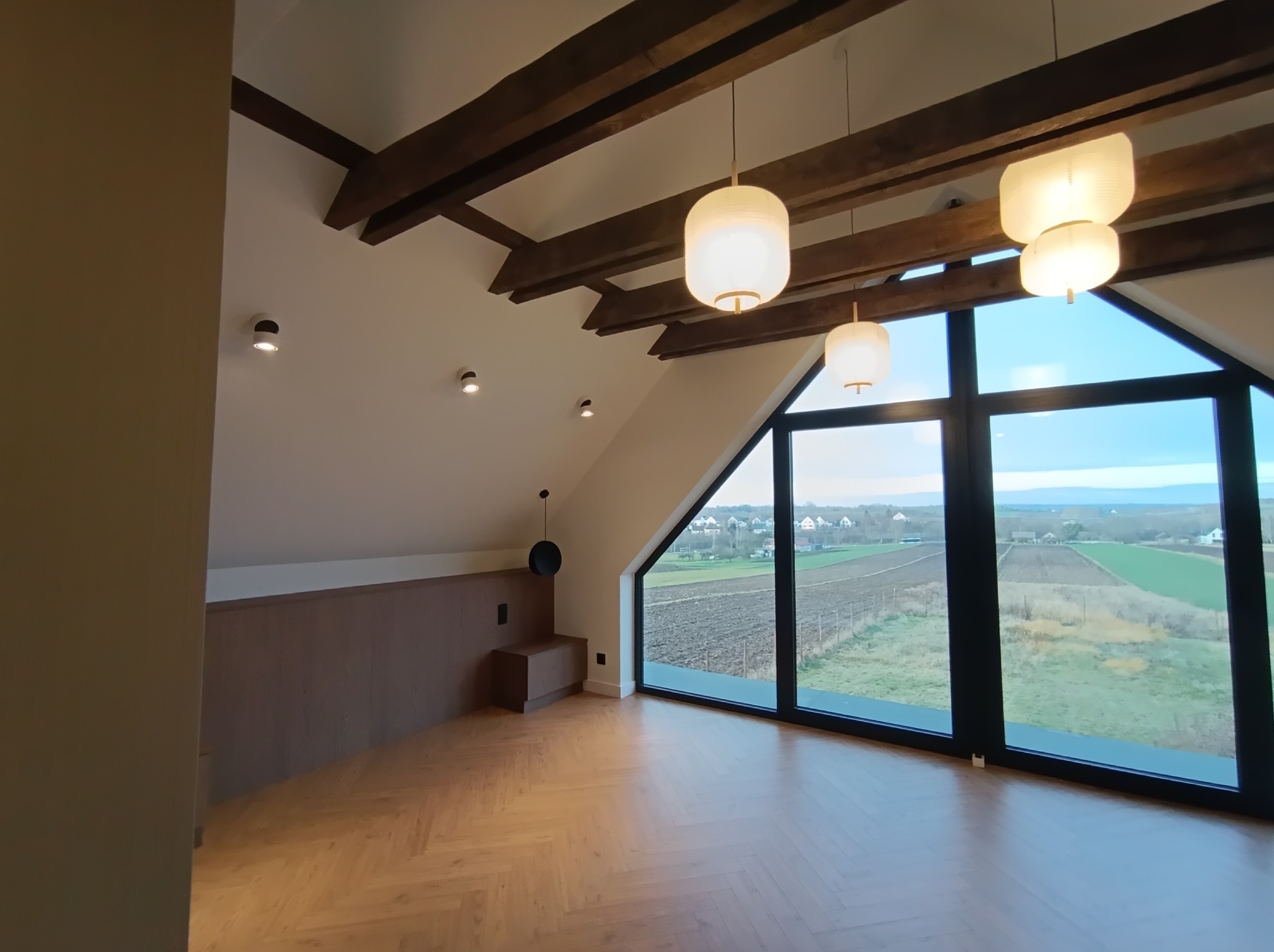 Jasne poddasze z parkietem w jodełkę, drewnianymi belkami i lampami w stylu skandynawskim. Duże okno z widokiem na krajobraz.