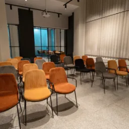 Klimatyzowana sala szkoleniowa z rzędami krzeseł w odcieniach brązu, szarości i czerni. Na ścianie projektor, w tle przeszklona ściana.