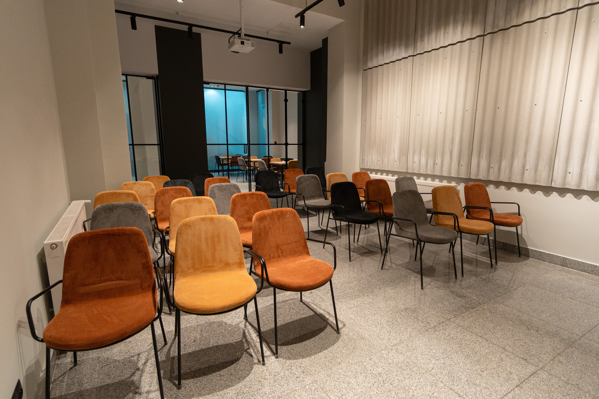 Klimatyzowana sala szkoleniowa z rzędami krzeseł w odcieniach brązu, szarości i czerni. Na ścianie projektor, w tle przeszklona ściana.