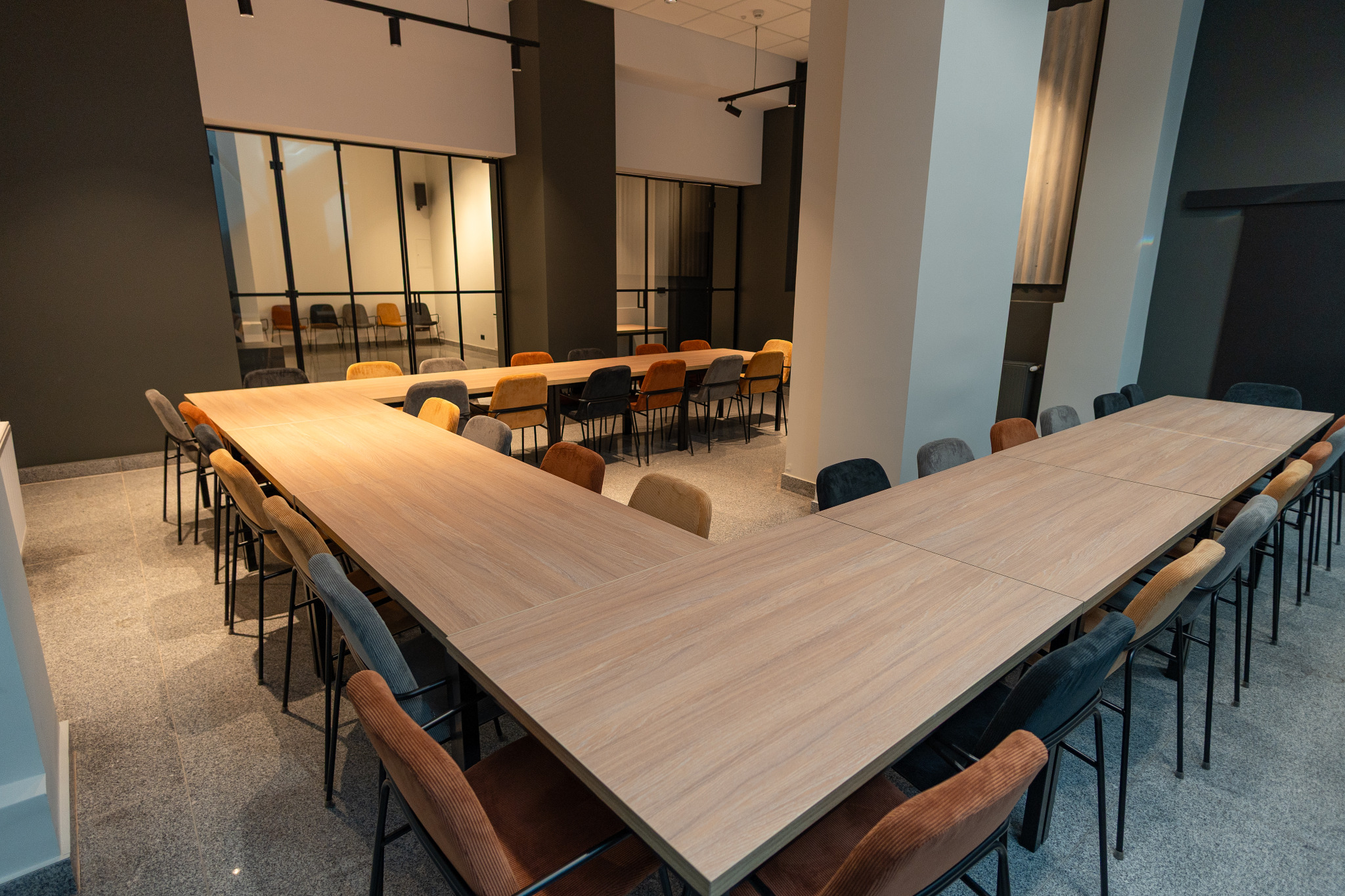 Sala konferencyjna z długim stołem w kształcie litery U i krzesłami w odcieniach szarości i brązu. Nowoczesne wnętrze z przeszkleniami i ciemnymi akcentami.