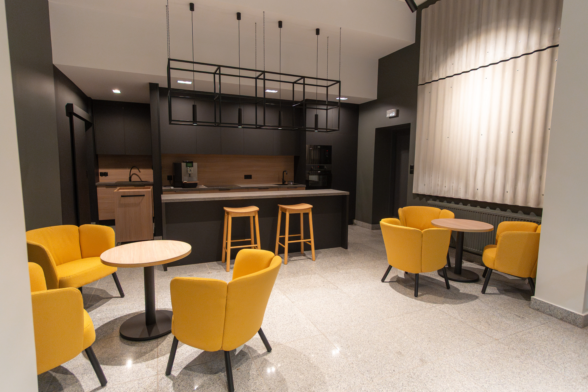 Nowoczesne wnętrze biurowe: żółte fotele klubowe przy okrągłych stolikach, minimalistyczna kuchnia z drewnianymi hokerami i designerską lampą.