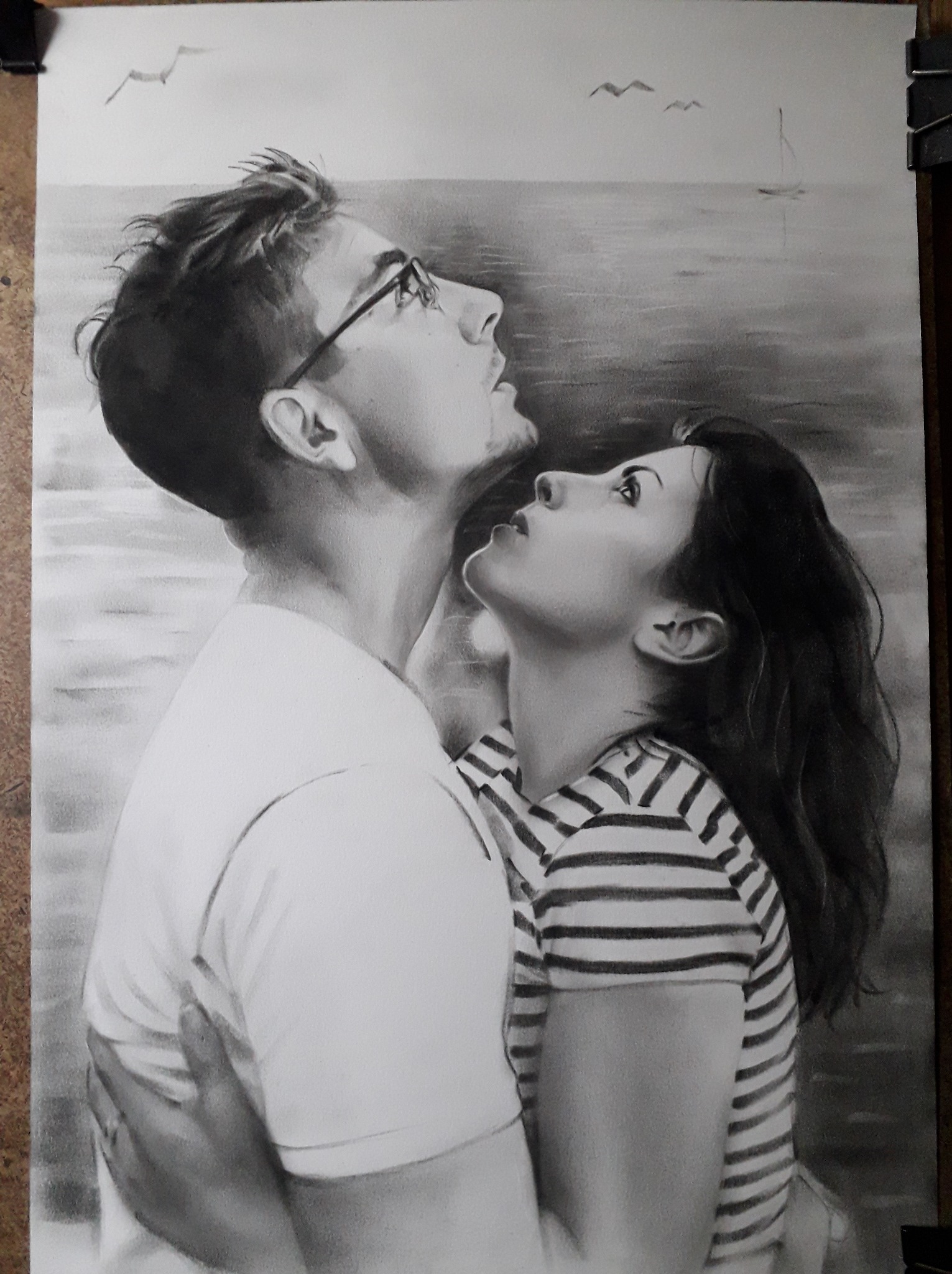 Czarno-biały portret pary spoglądającej w górę, na tle morza z żaglówką i mewami. Mężczyzna w okularach obejmuje kobietę w bluzce w paski. Rysunek wykonany ołówkiem.