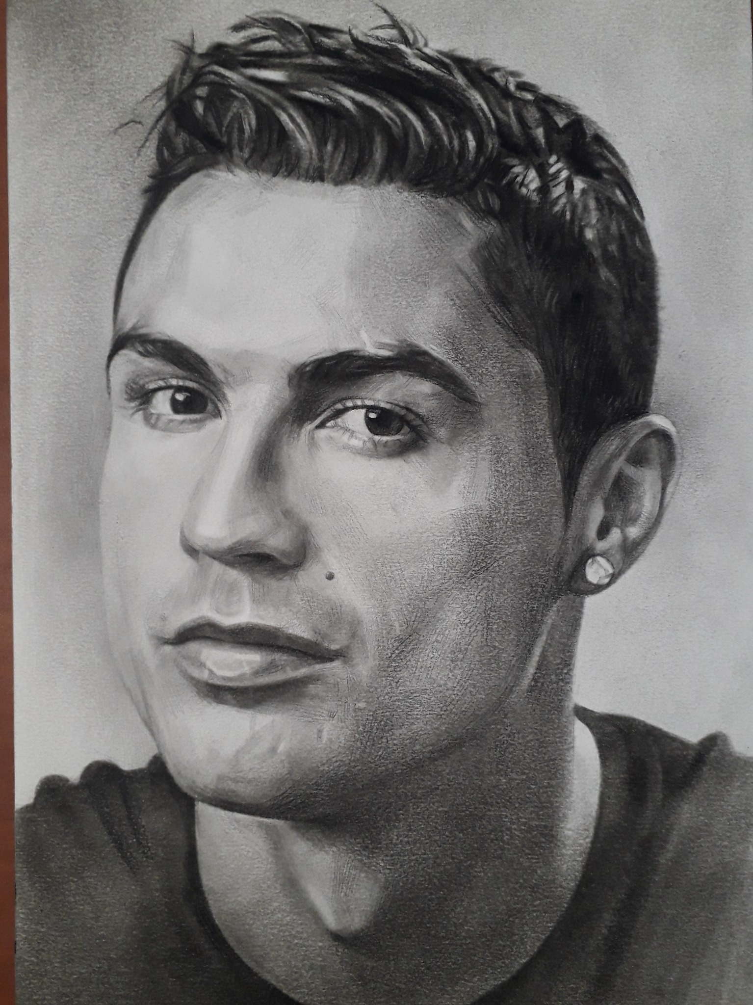 Portret Ronaldo narysowany ołówkiem, z widocznymi detalami twarzy i włosów, na szarym tle. Widoczny kolczyk w uchu. Realistyczny styl.