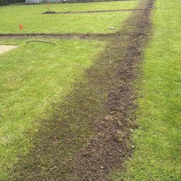 Gaja Gardens Joanna Lalka - Wyżłobione bruzdy w trawniku, prawdopodobnie pod instalację nawadniającą lub przewody elektryczne. W tle domek narzędziowy i rząd tuj.