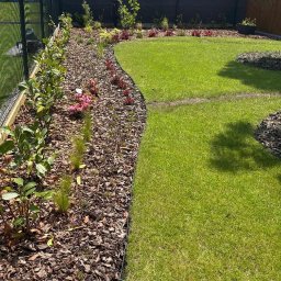 Gaja Gardens Joanna Lalka - Zadbany ogród z równo przystrzyżonym trawnikiem i rabatami wyłożonymi korą. Widoczne świeżo posadzone krzewy i kwiaty. Estetyczne obrzeża oddzielają trawnik od rabat.