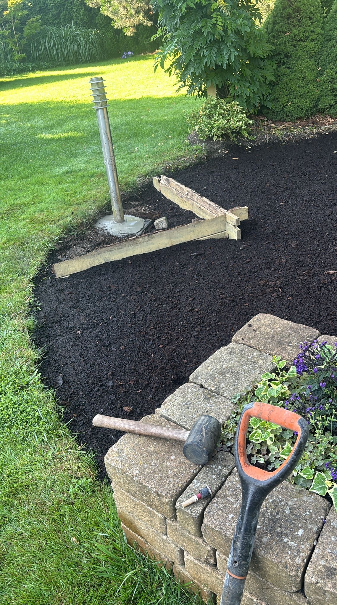 Przygotowanie terenu pod rabatę: świeżo rozsypana kora, obrzeże z kostki, narzędzia ogrodnicze (szpadel, młotek) i lampa ogrodowa w tle.