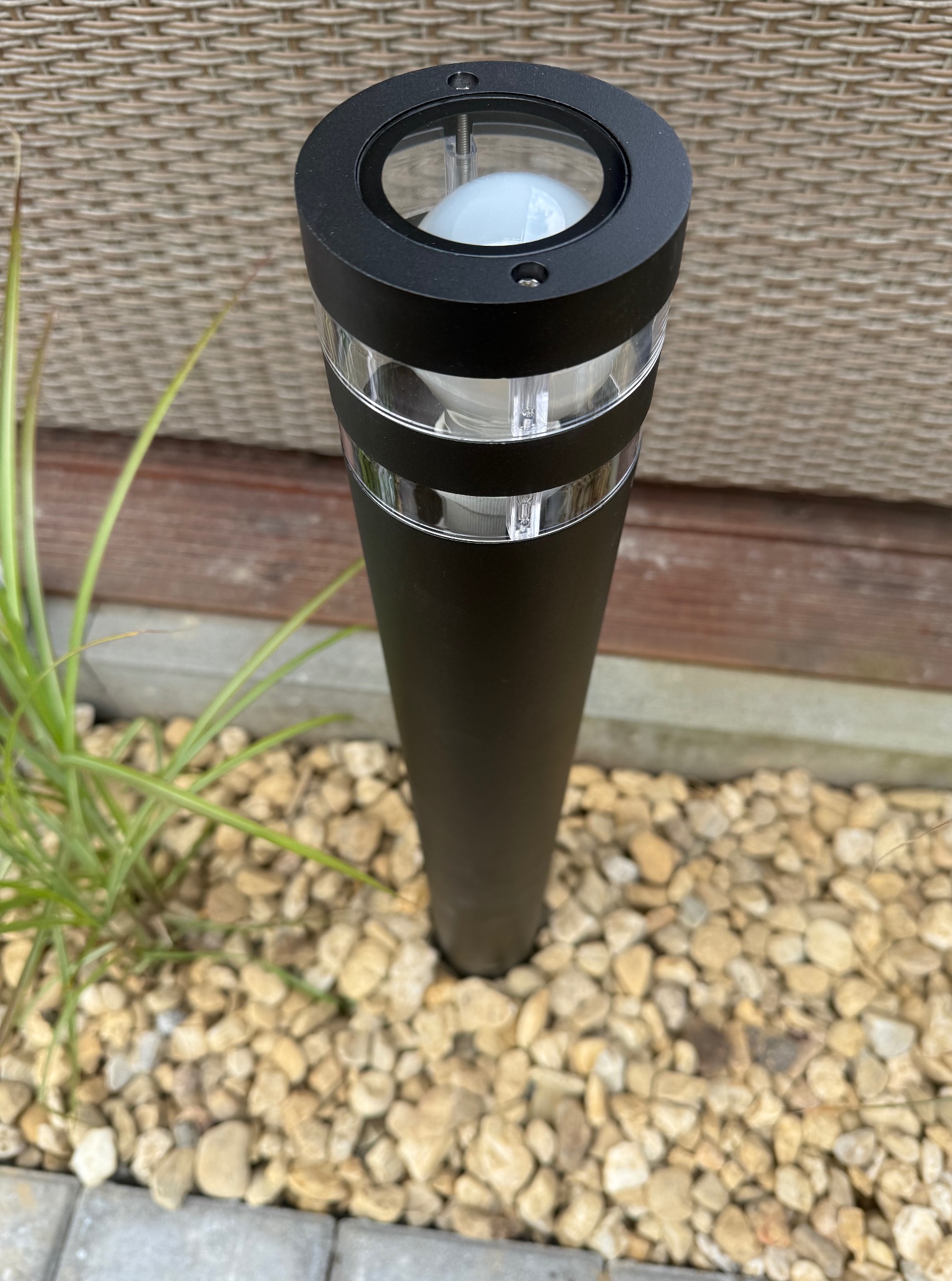 Czarna lampa ogrodowa stojąca na żwirze, oświetlająca przestrzeń. Minimalistyczny design, z elementami przezroczystymi. W tle ściana z brązowego rattanu.