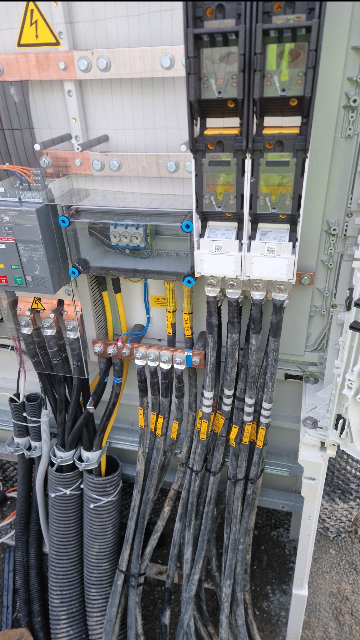 Rozdzielnica elektryczna z podłączonymi kablami oznaczonymi żółtymi etykietami. Widoczne bezpieczniki, przewody i listwy zaciskowe. Instalacja w szarej obudowie.