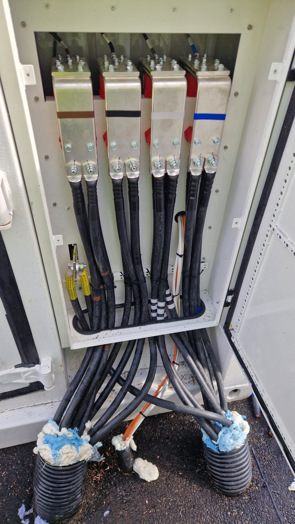 Rozdzielnica elektryczna z grubymi kablami zabezpieczonymi pianką montażową, z metalowymi złączkami oznaczonymi kolorami. Widok z góry na instalację.