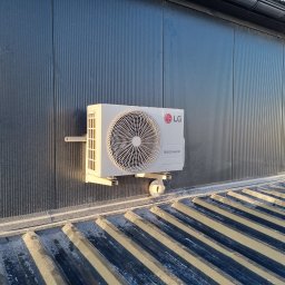 VG sp. z o.o. - Zamontowana jednostka zewnętrzna klimatyzacji LG Dual Inverter na ciemnej, metalowej ścianie budynku z widocznym fragmentem dachu. Ujęcie z dołu.