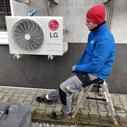 VG sp. z o.o. - Instalator siedzi na drabinie obok zamontowanych klimatyzatorów LG na szarej ścianie budynku. Widoczne przewody i elementy montażowe. Ujęcie z dołu.