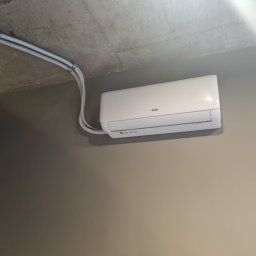 VG sp. z o.o. - Biały klimatyzator AUX zamontowany na szarej ścianie, widoczne rury instalacyjne przymocowane do sufitu. Minimalistyczny design.