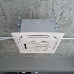 VG sp. z o.o. - Nowoczesna, biała klimatyzacja kasetonowa AUX zamontowana pod surowym, betonowym sufitem. Widoczne metalowe pręty mocujące i biała instalacja elektryczna.