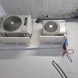 VG sp. z o.o. - Dwa białe klimatyzatory AUX zamontowane na ścianie, z widocznymi przewodami instalacyjnymi i rurami wentylacyjnymi pod sufitem. Widok z dołu.